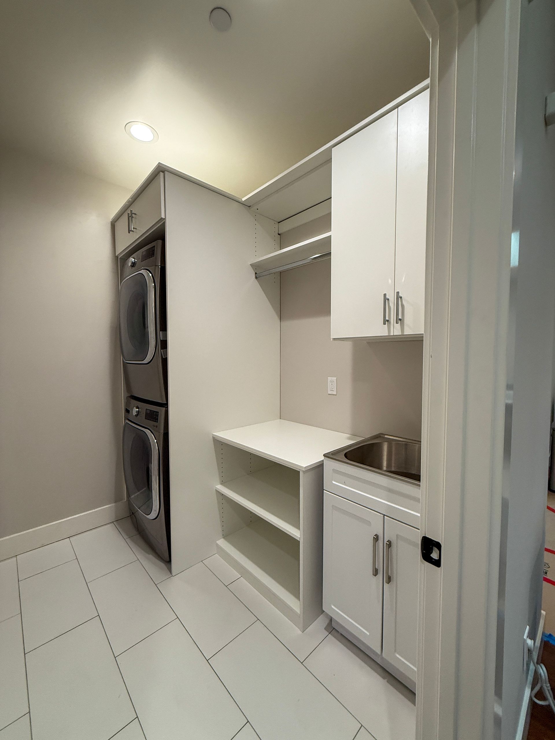 A white laundry room featuring a stacked washer and dryer, open shelving, a sink with cabinets, and a tile floor.