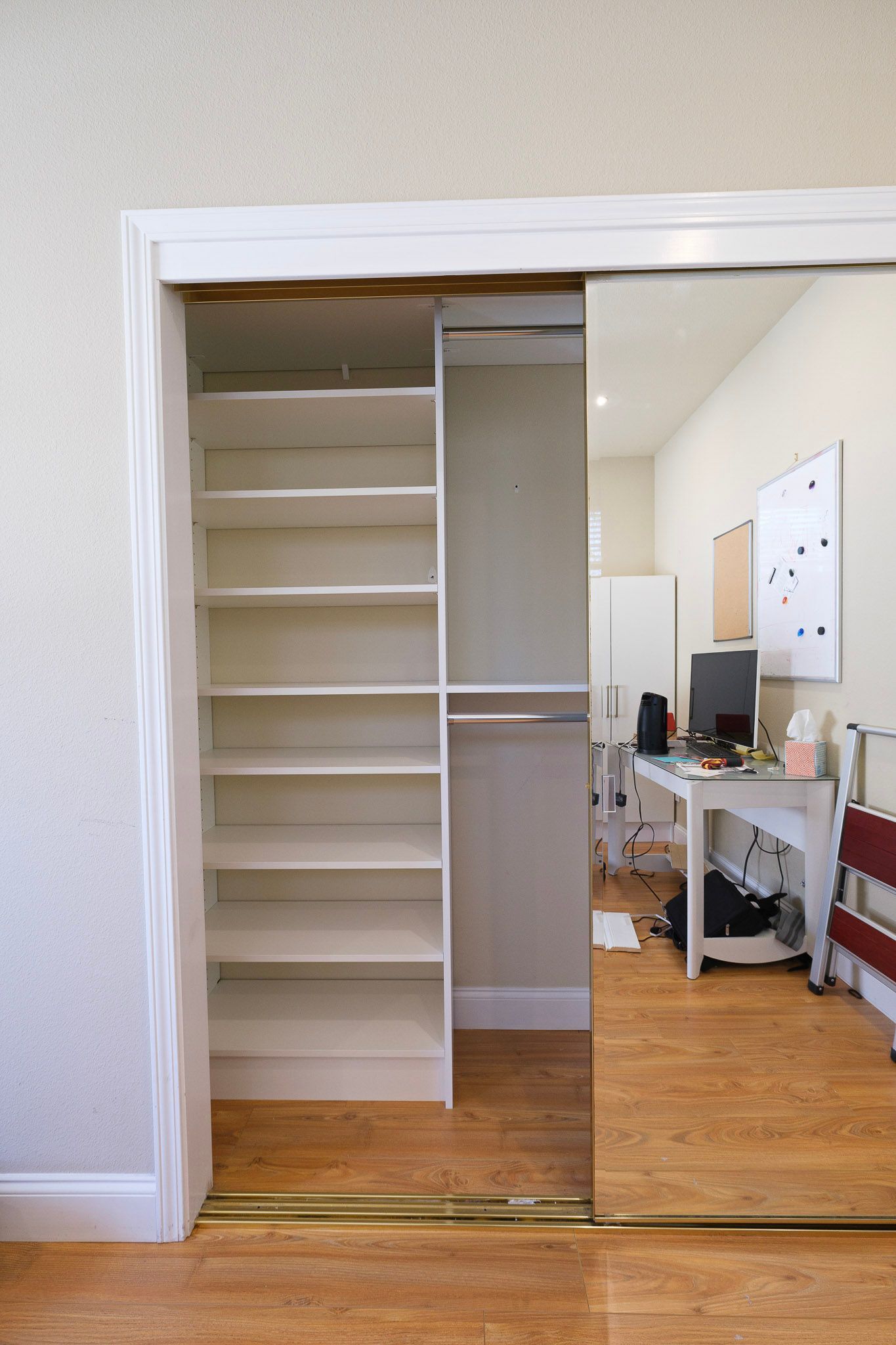 A built-in closet with white shelving and a hanging rod, reflected in sliding mirror doors with a view of a desk beyond.