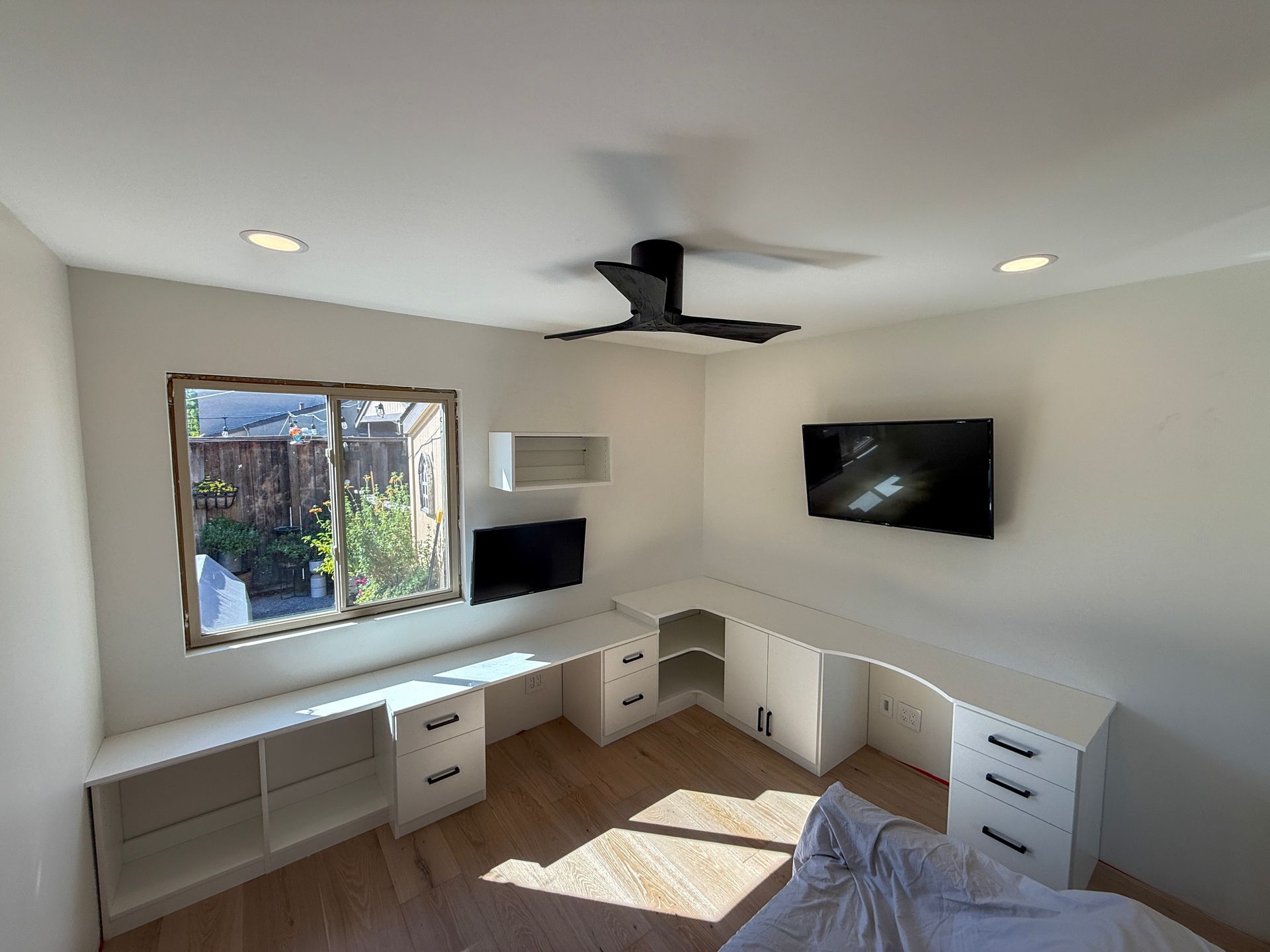 A room with white, L-shaped built-in desks, drawers, and shelving, featuring two wall-mounted televisions and a ceiling fan.