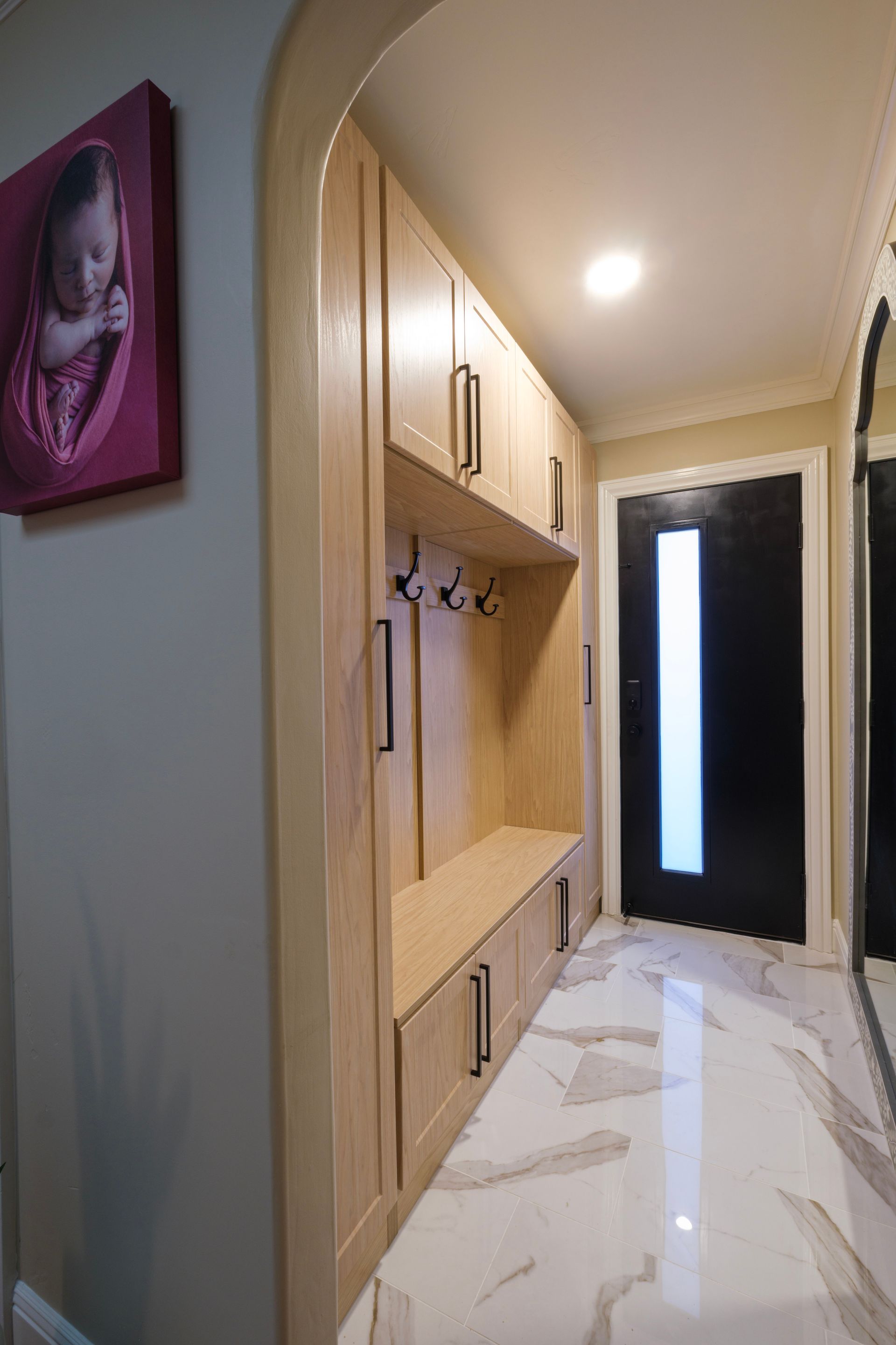 A light wood entryway mudroom with cabinets, coat hooks, and a bench, next to a black door in a white tiled hallway.