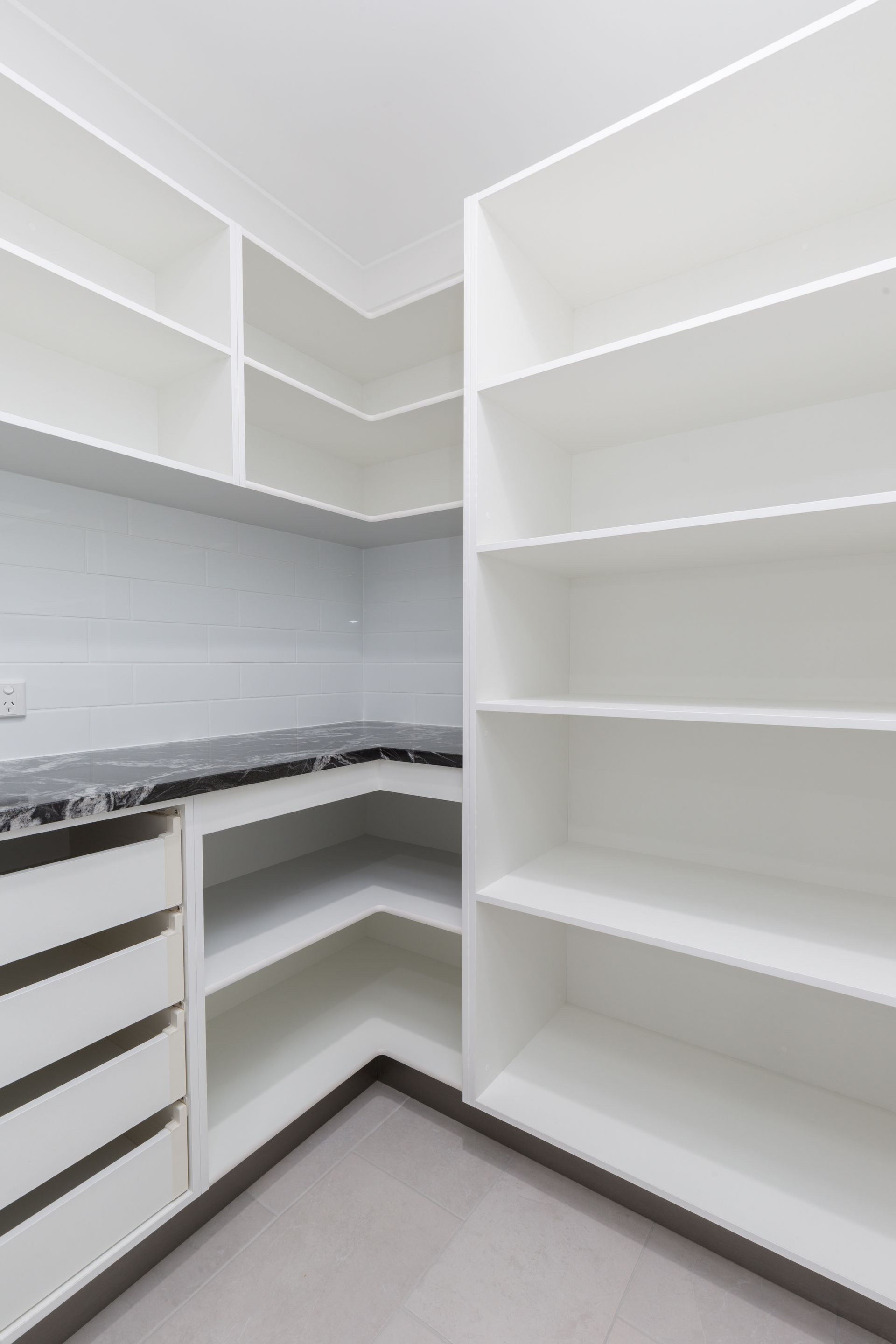 A white pantry with L-shaped shelving, drawers, and a dark marble-look countertop against a white tiled wall.