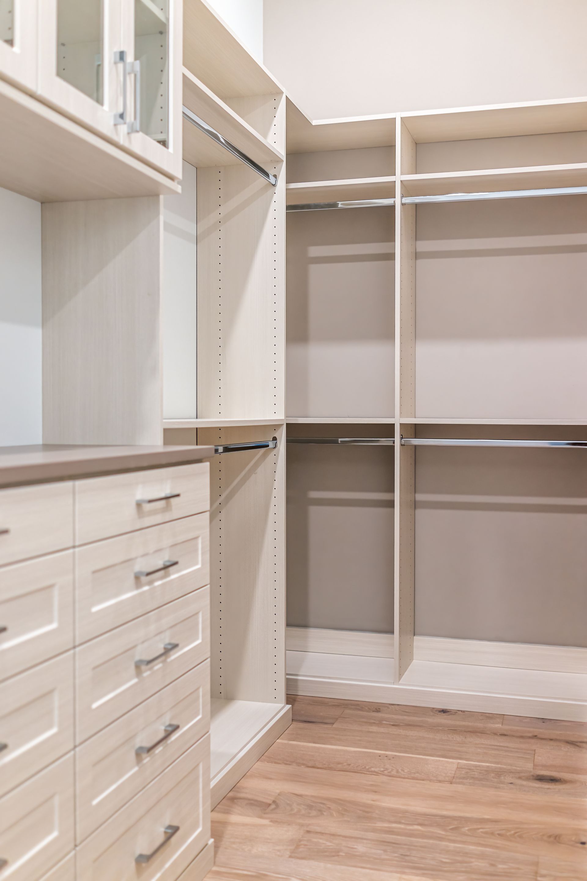 Empty walk-in closet with cream-colored built-in shelves, drawers, hanging rods, and light wood flooring.