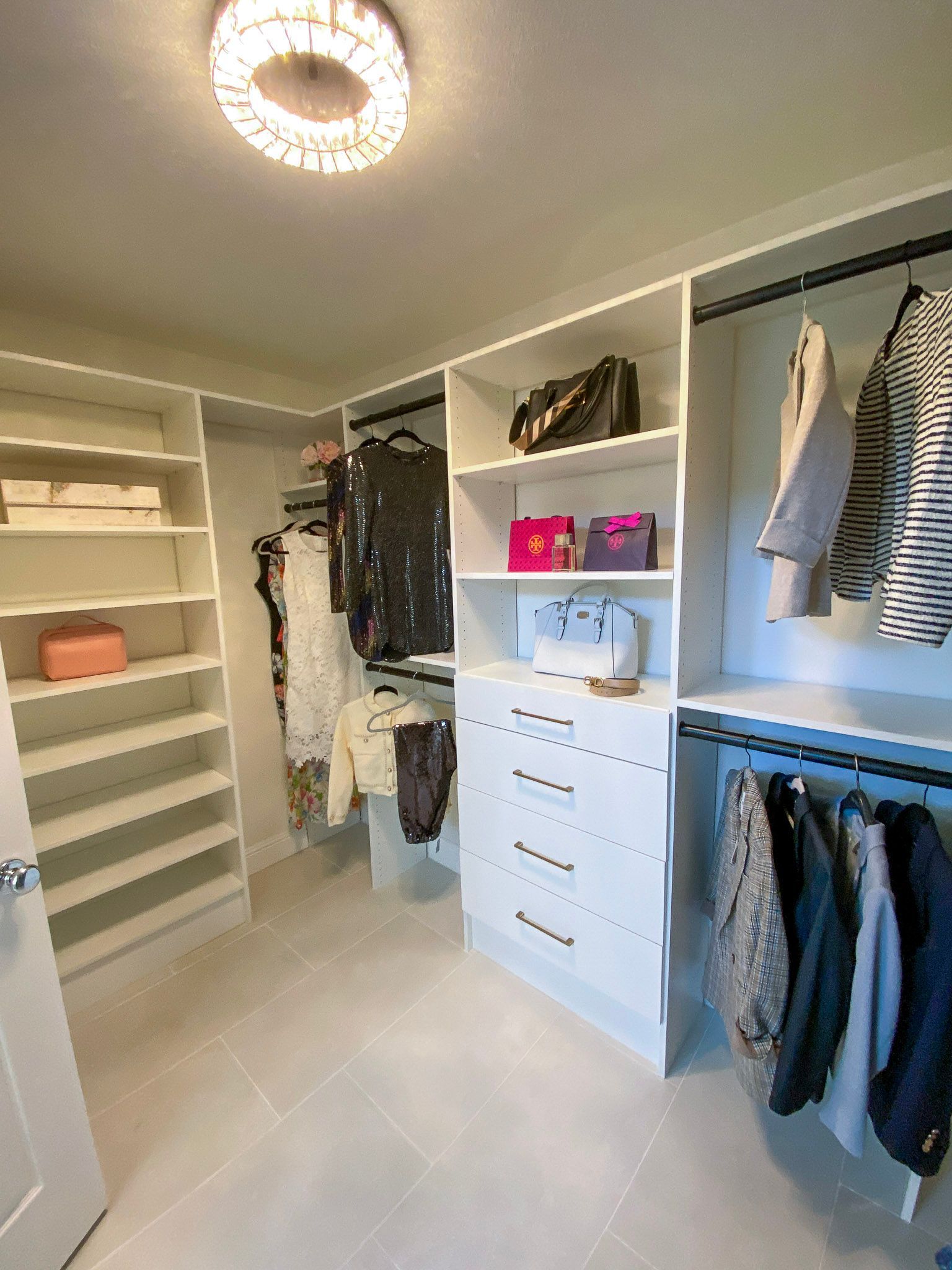 A bright, modern walk-in closet with white shelving, hanging clothes, drawers, and an overhead crystal light fixture.
