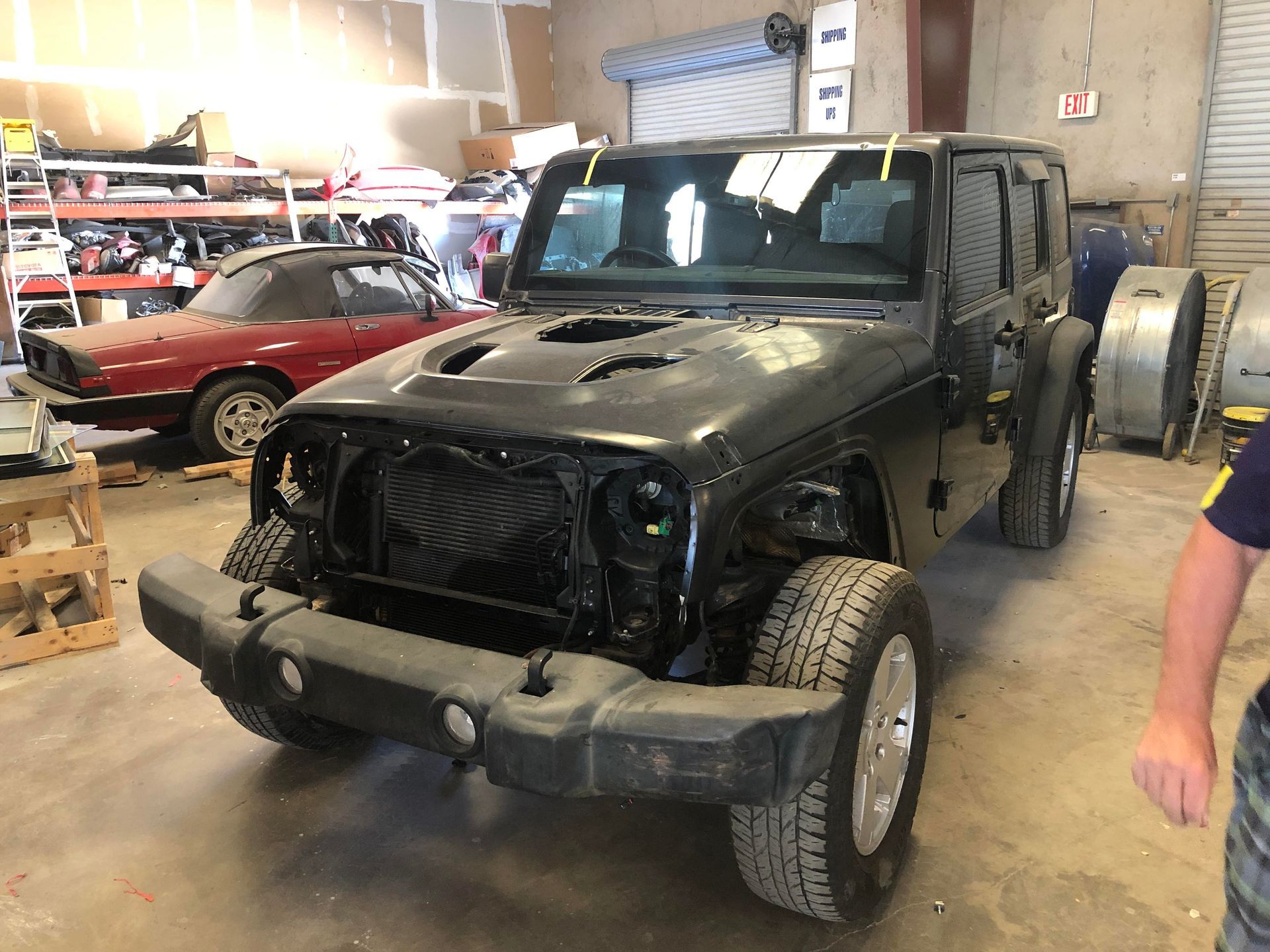 Black Jeep Wrangler with missing front end in a garage. Red convertible in the background.