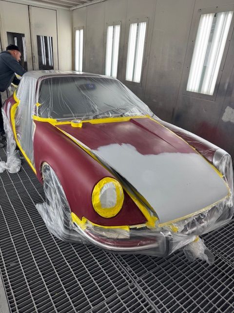 Car in a paint booth, partially painted red and gray, covered in plastic. Person in the background.