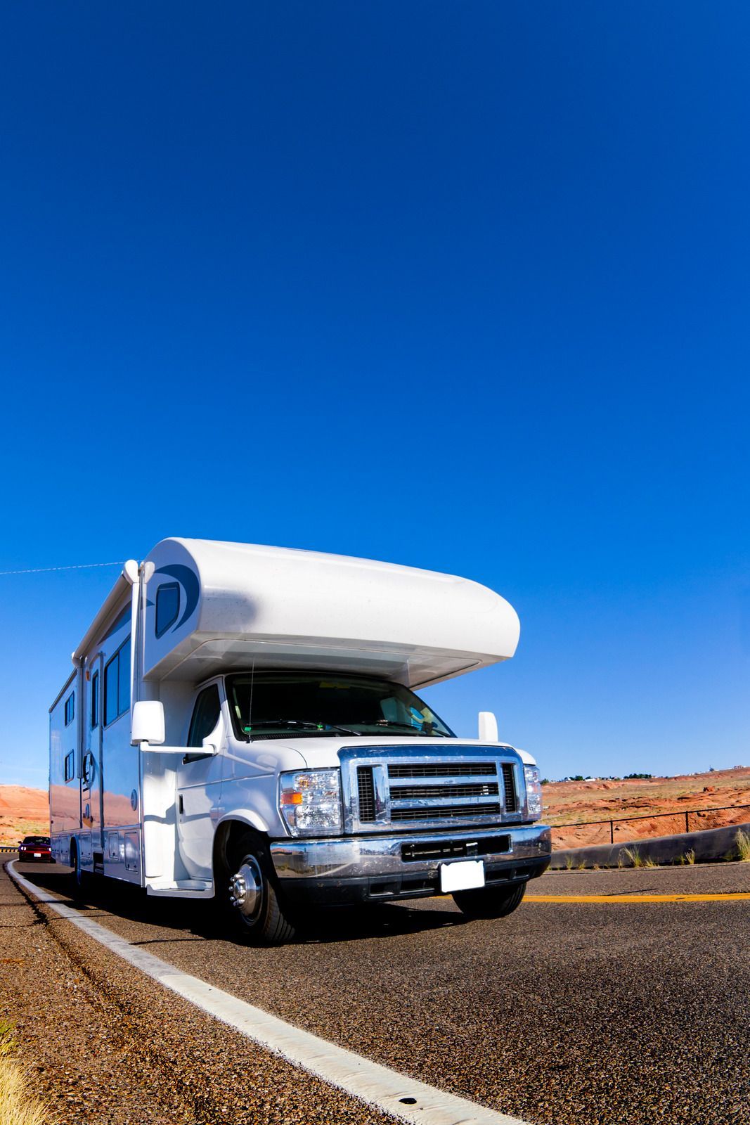 White RV on a desert road with red rocks and a clear blue sky.