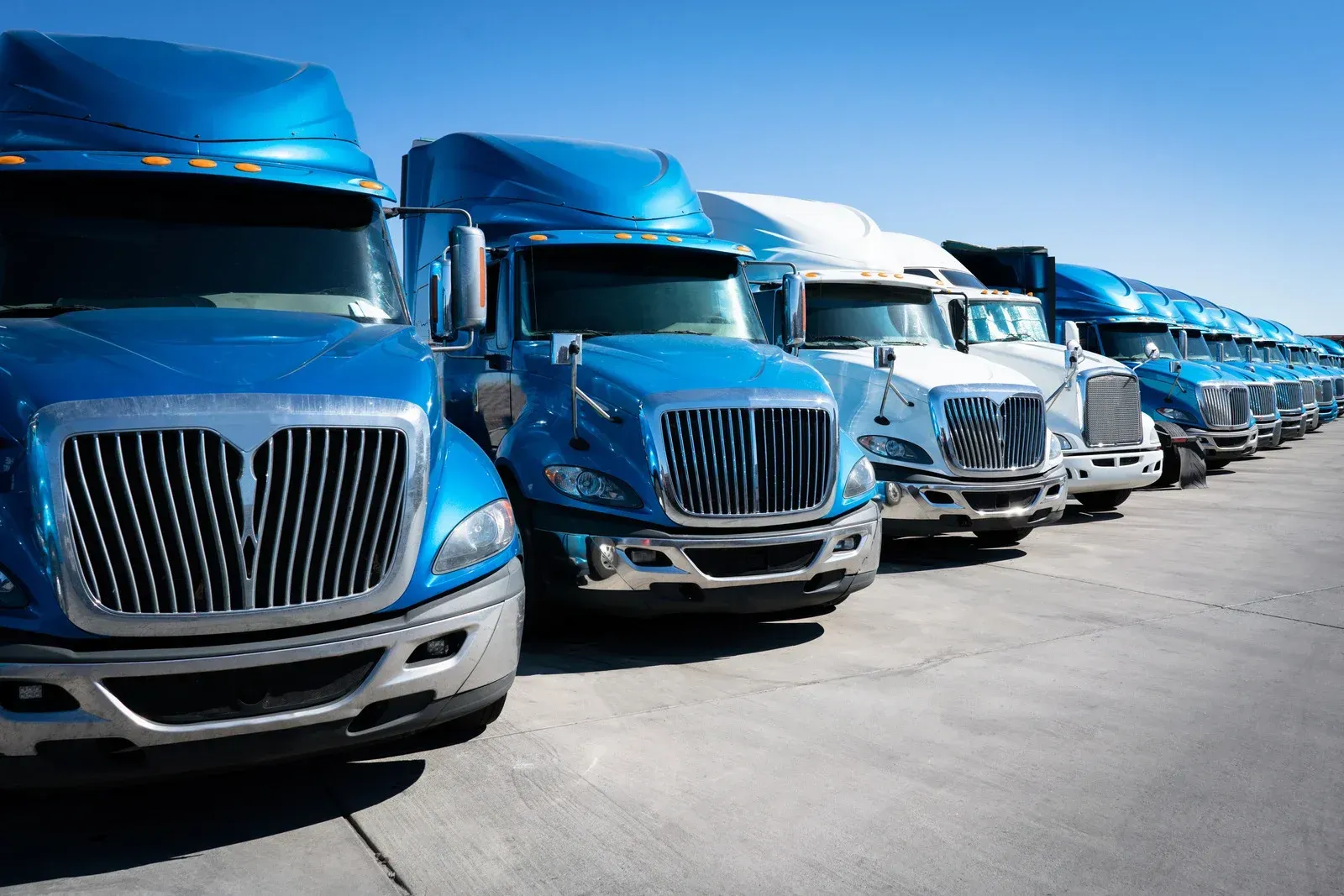 Row of blue and white semi-trucks parked on a paved lot under a clear blue sky.