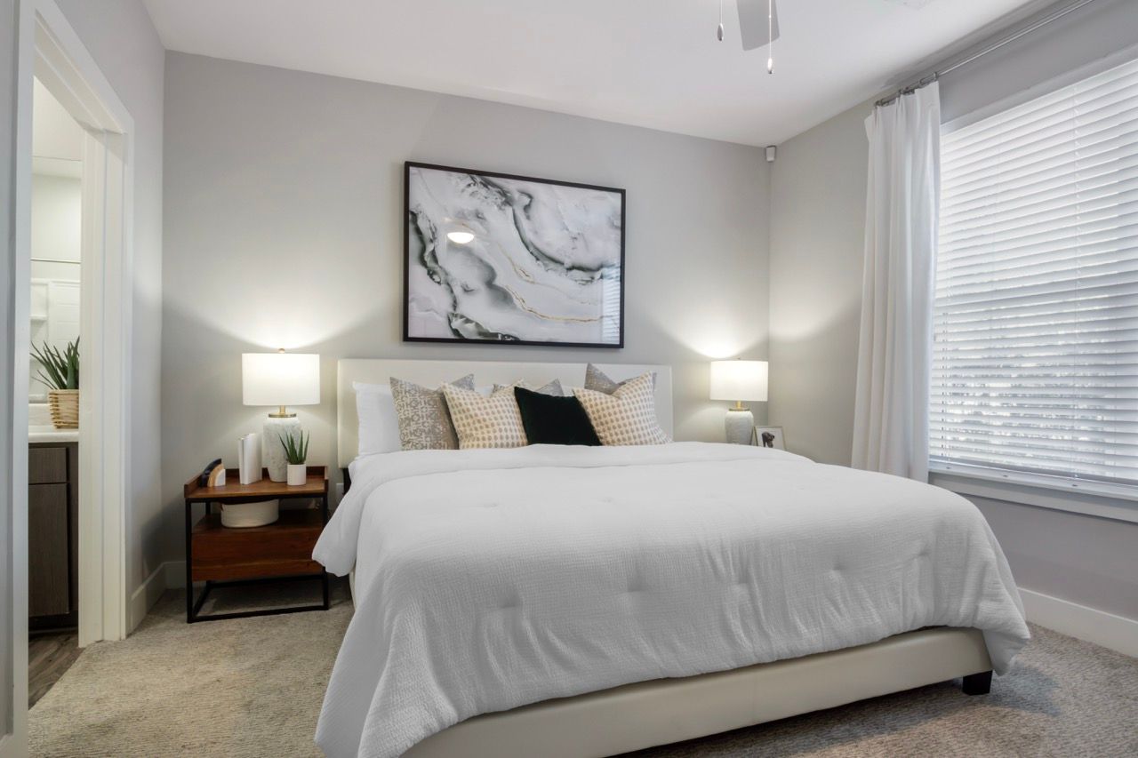 Bright bedroom with a white bed, two nightstands, lamps, and a large window with blinds.