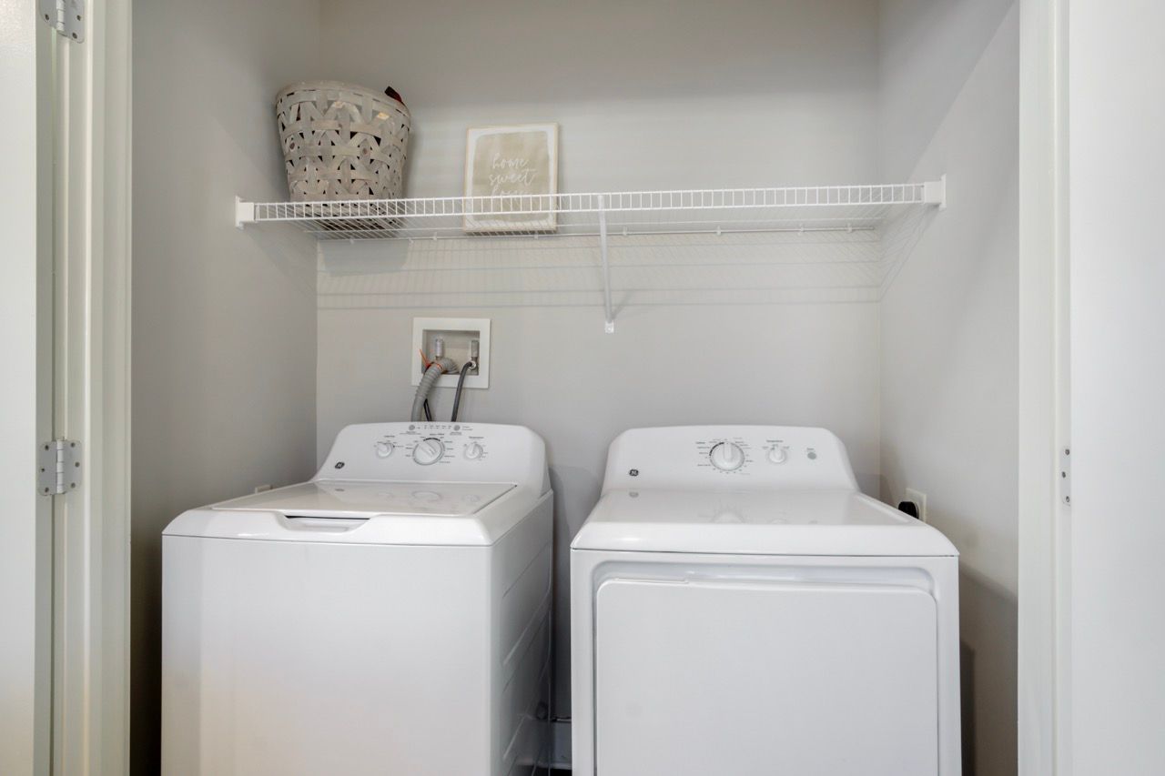 Laundry closet with a washer and dryer side by side, white walls, and a wire shelf above.