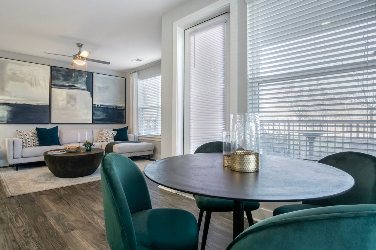 Open-concept living and dining area in an apartment with large windows and teal chairs.