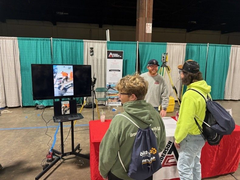 Three people stand at a trade show booth, looking at a demonstration on a monitor.