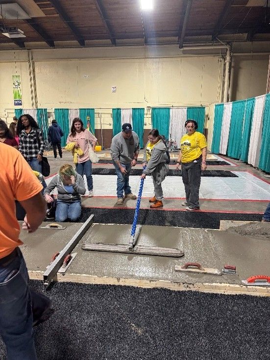 A group of people works in a large, indoor facility, using tools to smooth wet concrete over a dark, textured base.