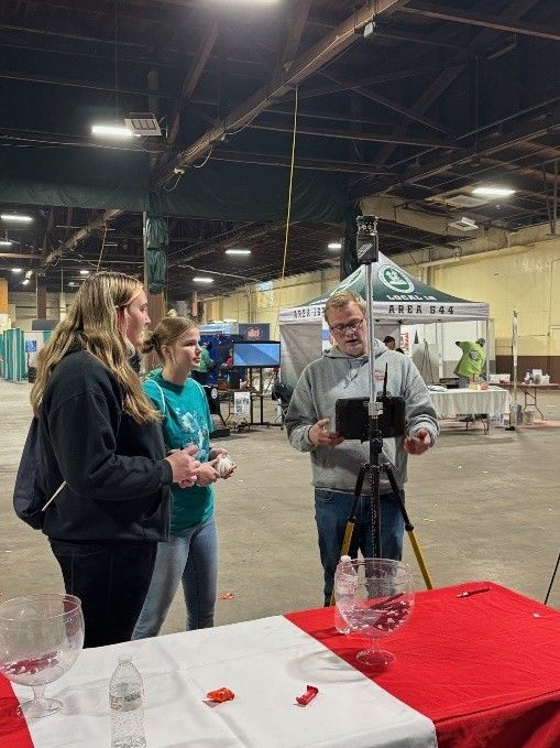 Three people stand around a table with a tripod-mounted camera, demonstrating equipment in a large indoor venue.
