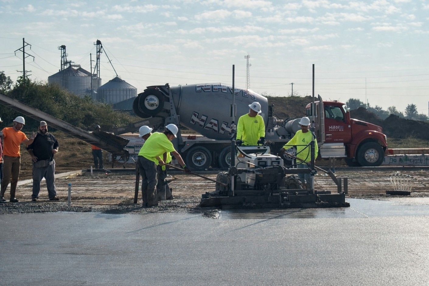 Construction workers guide a concrete screed machine while a cement truck pours mix onto a large foundation site.