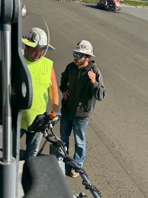 Two workers in hard hats and safety gear stand on a paved surface, discussing work while using a handheld tablet device.
