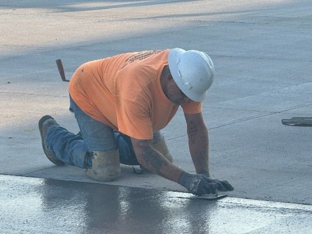 A construction worker in an orange shirt and white hard hat kneels, smoothing wet concrete with a hand tool.