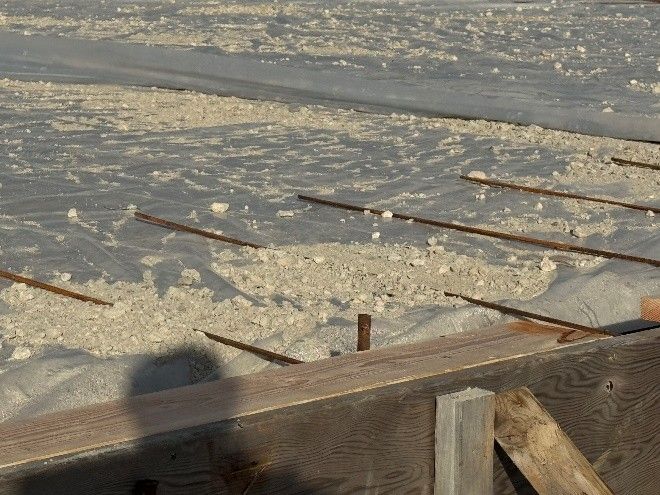 Construction site showing a wooden formwork frame with rebar laid over a vapor barrier and gravel base for a concrete pour.