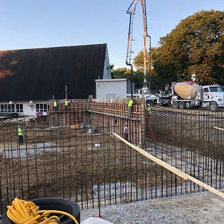 Construction site with workers, rebar, and concrete being poured from a truck. Church building in the background.