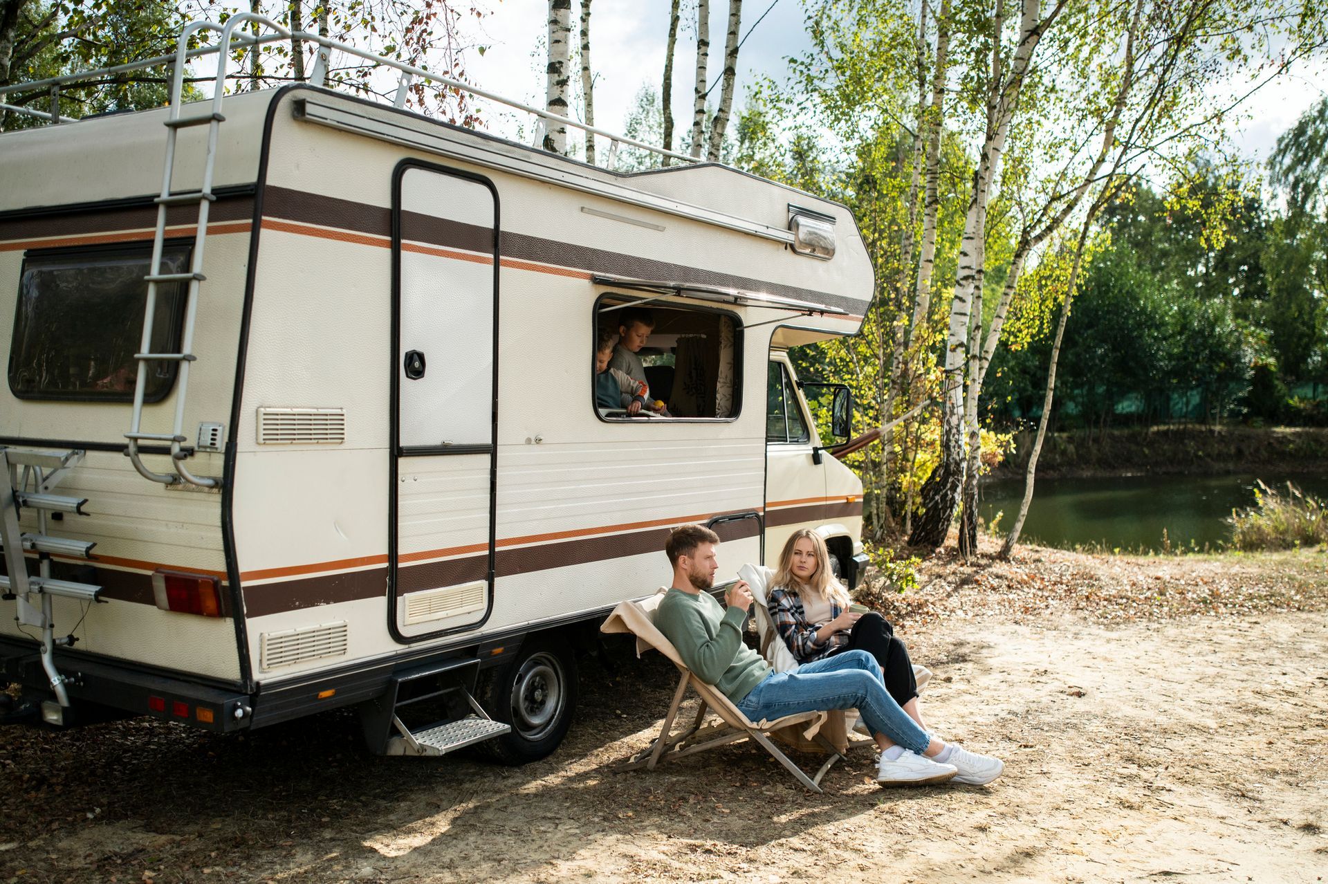 Camper parked by a lake with two people relaxing in chairs outdoors.