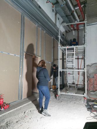 Personas trabajando en la construcción dentro de un edificio; andamios y paneles de yeso en su lugar.