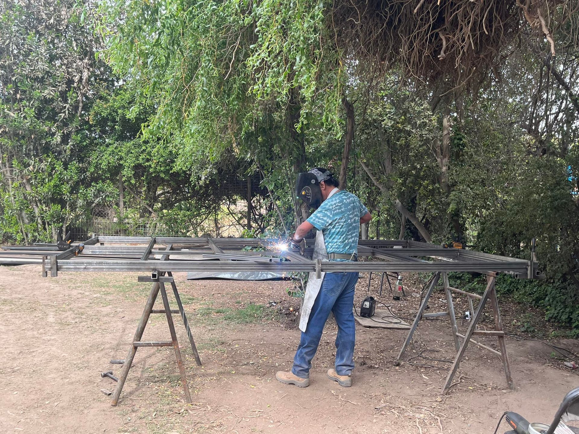 Hombre soldando un marco de metal al aire libre, vistiendo un casco y un delantal de soldador.