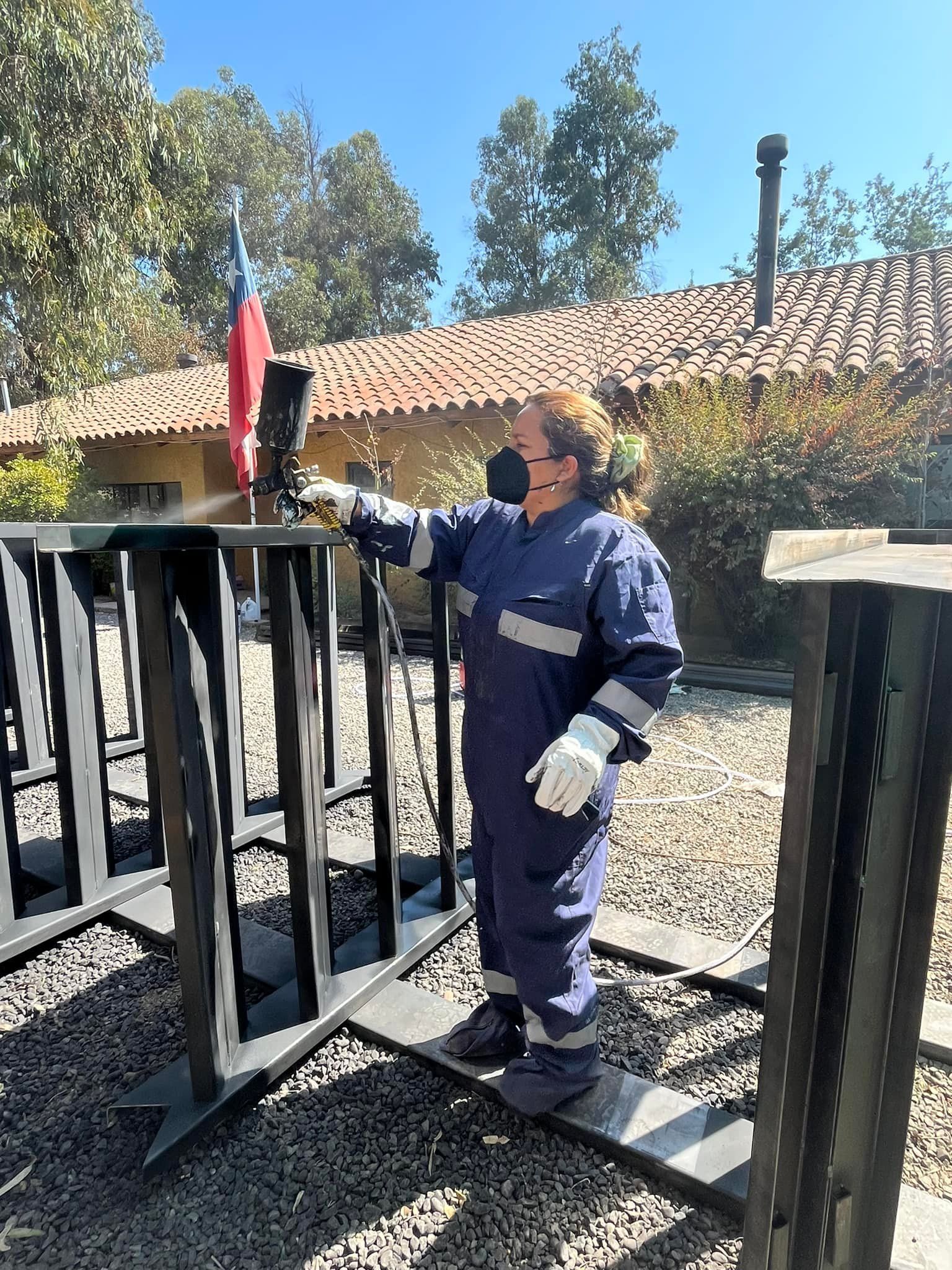 Una mujer con un mono azul rocía pintura sobre una estructura de metal al aire libre, con la bandera chilena de fondo.