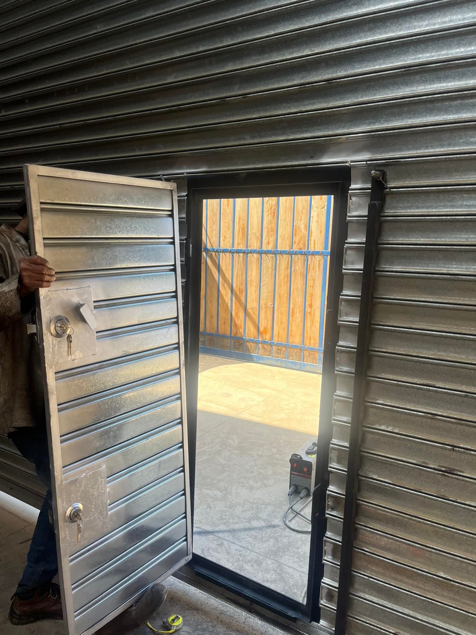 Persona que abre una puerta metálica resistente dentro de una estructura de metal corrugado, revelando un área al aire libre.