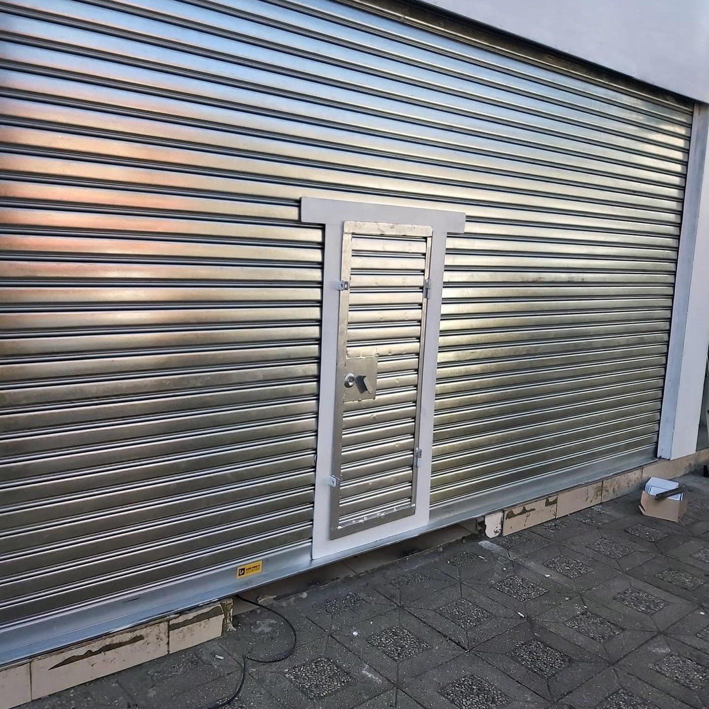 Puerta de seguridad enrollable de metal plateado con pequeña puerta de acceso, en el exterior del edificio.