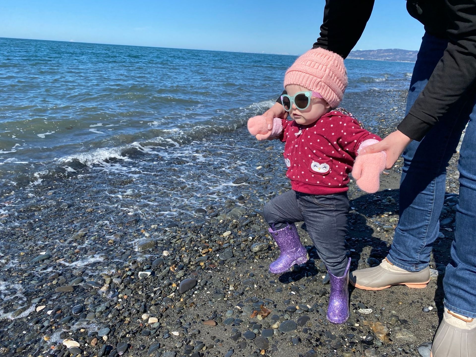 A little girl wearing sunglasses and purple boots is walking on the beach.