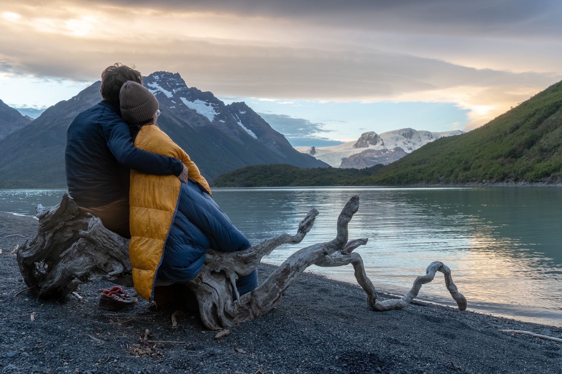 A couple in sleeping bags is sitting on a log by a lake.