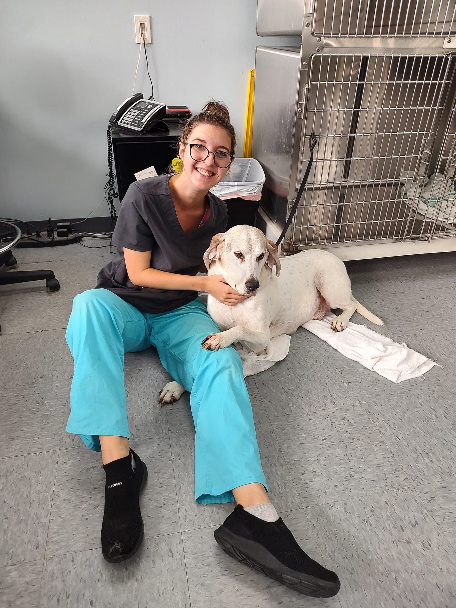 Woman in teal scrubs sits on the floor petting a white dog, inside a vet clinic.