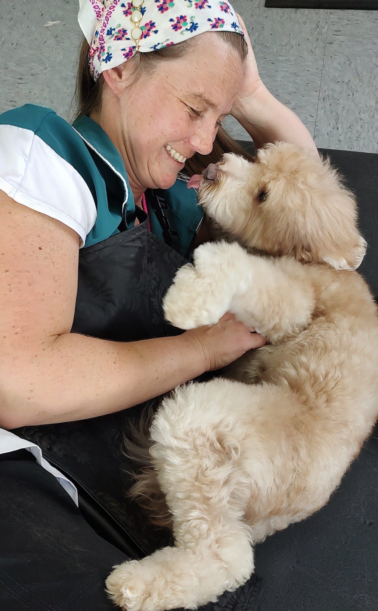 Woman with floral headscarf smiling, playfully interacting with a fluffy, light-colored dog on a dark surface.