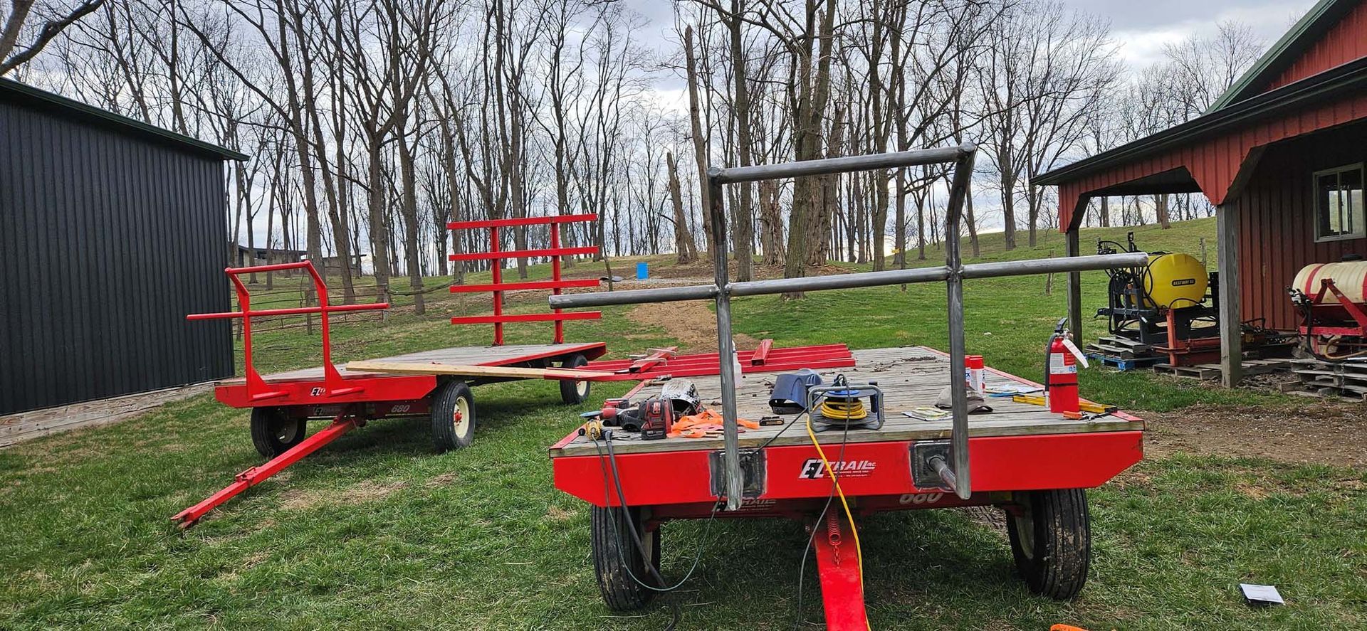 Two red trailers are parked in a grassy field in front of a barn.