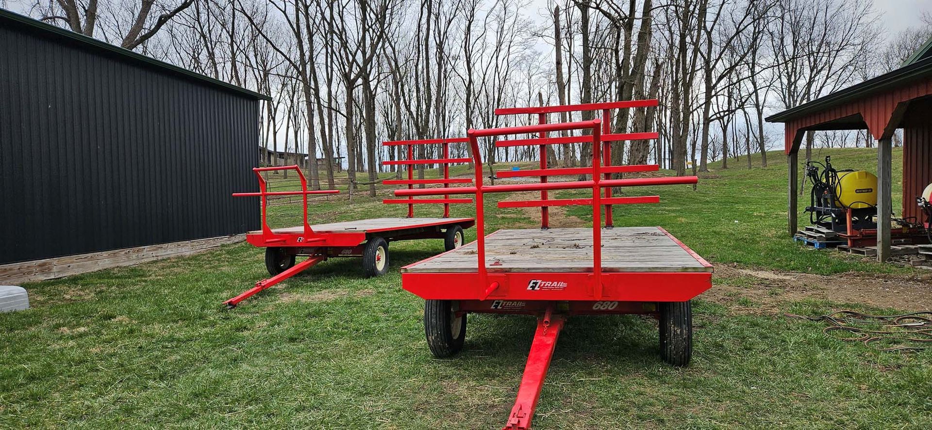 Two red trailers are parked in a grassy field in front of a shed.