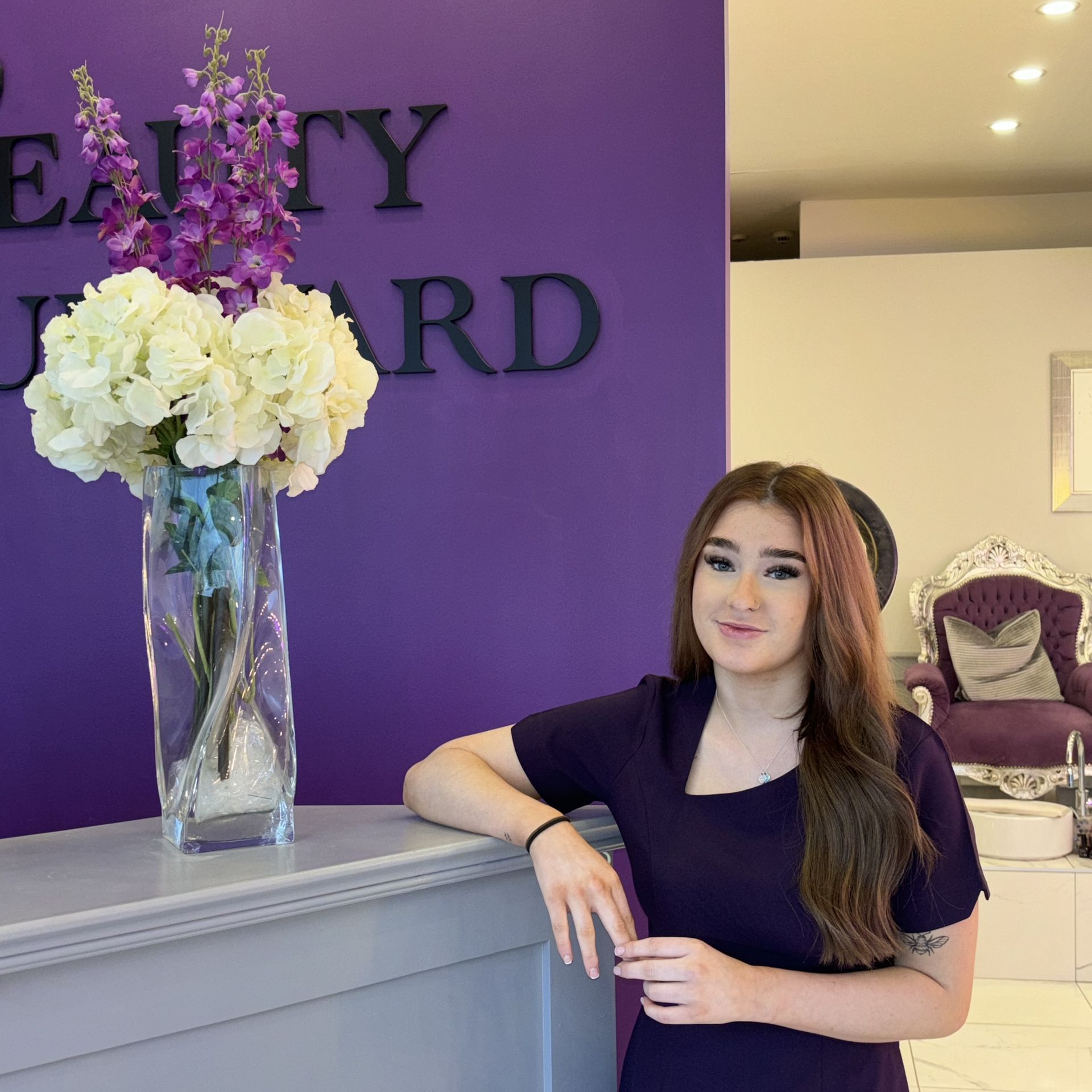 Woman with blonde hair leans on a salon reception desk in front of a purple wall with a business sign.