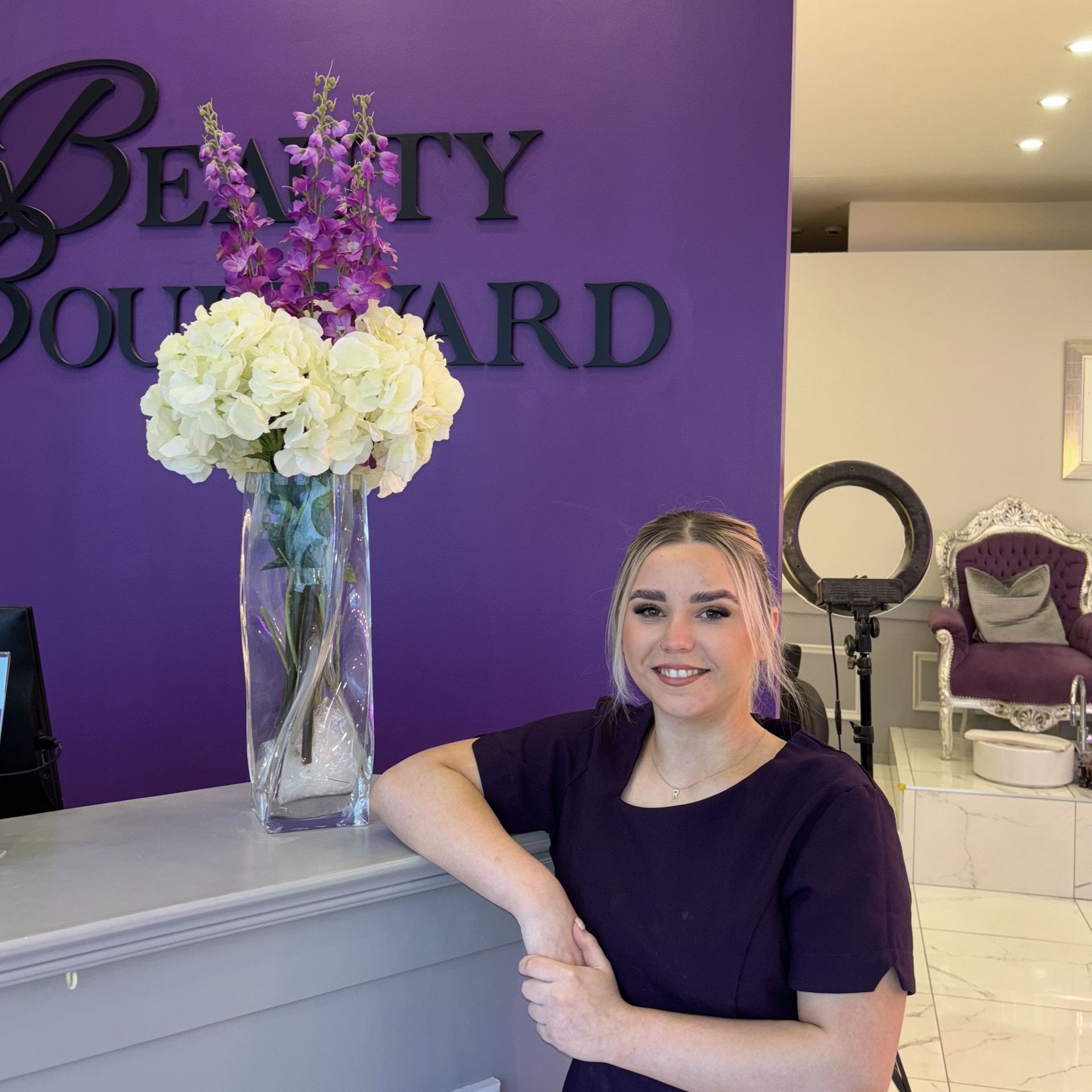 Woman with blonde hair leans on a salon reception desk in front of a purple wall with a business sign.
