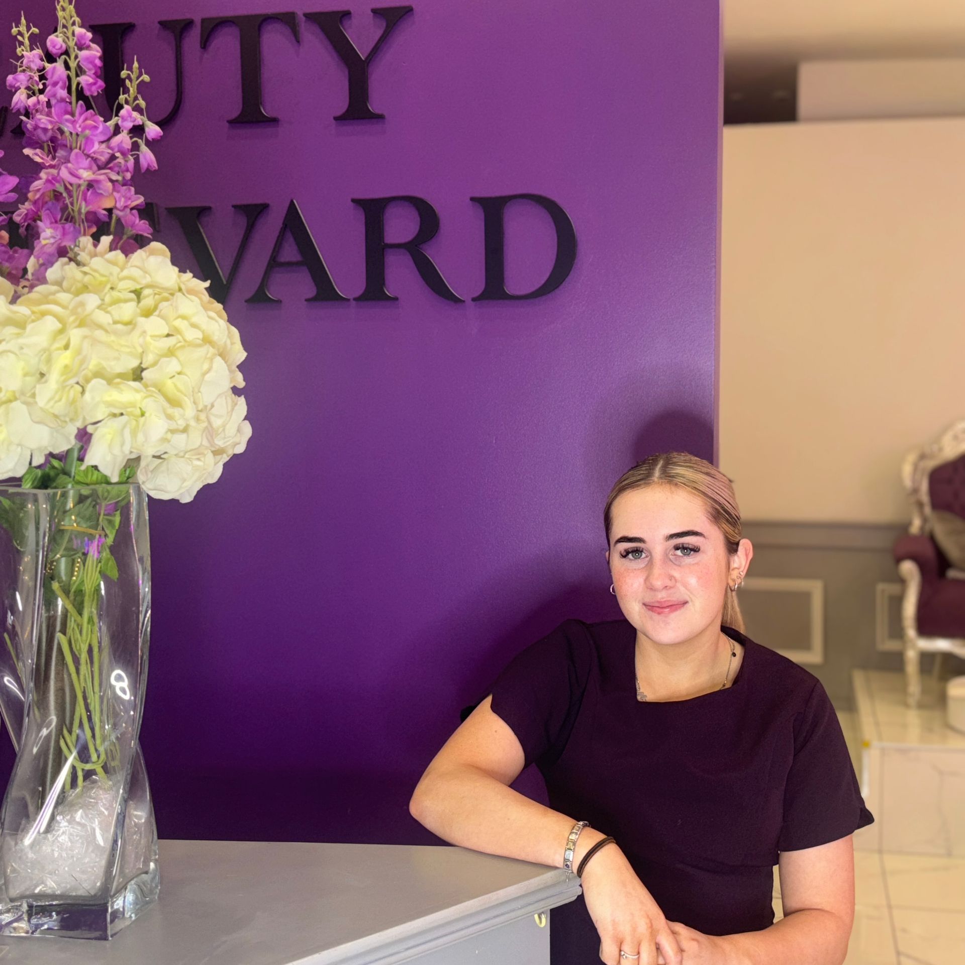 Woman with blonde hair leans on a salon reception desk in front of a purple wall with a business sign.