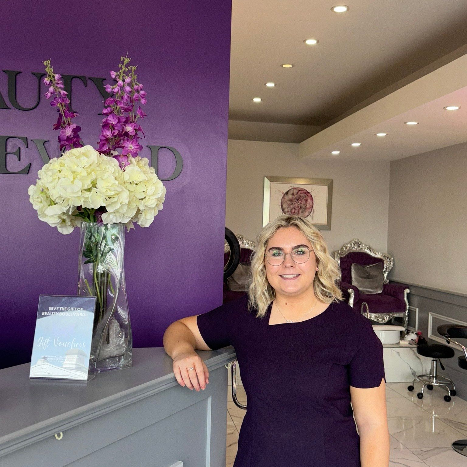Woman with blonde hair leans on a salon reception desk in front of a purple wall with a business sign.
