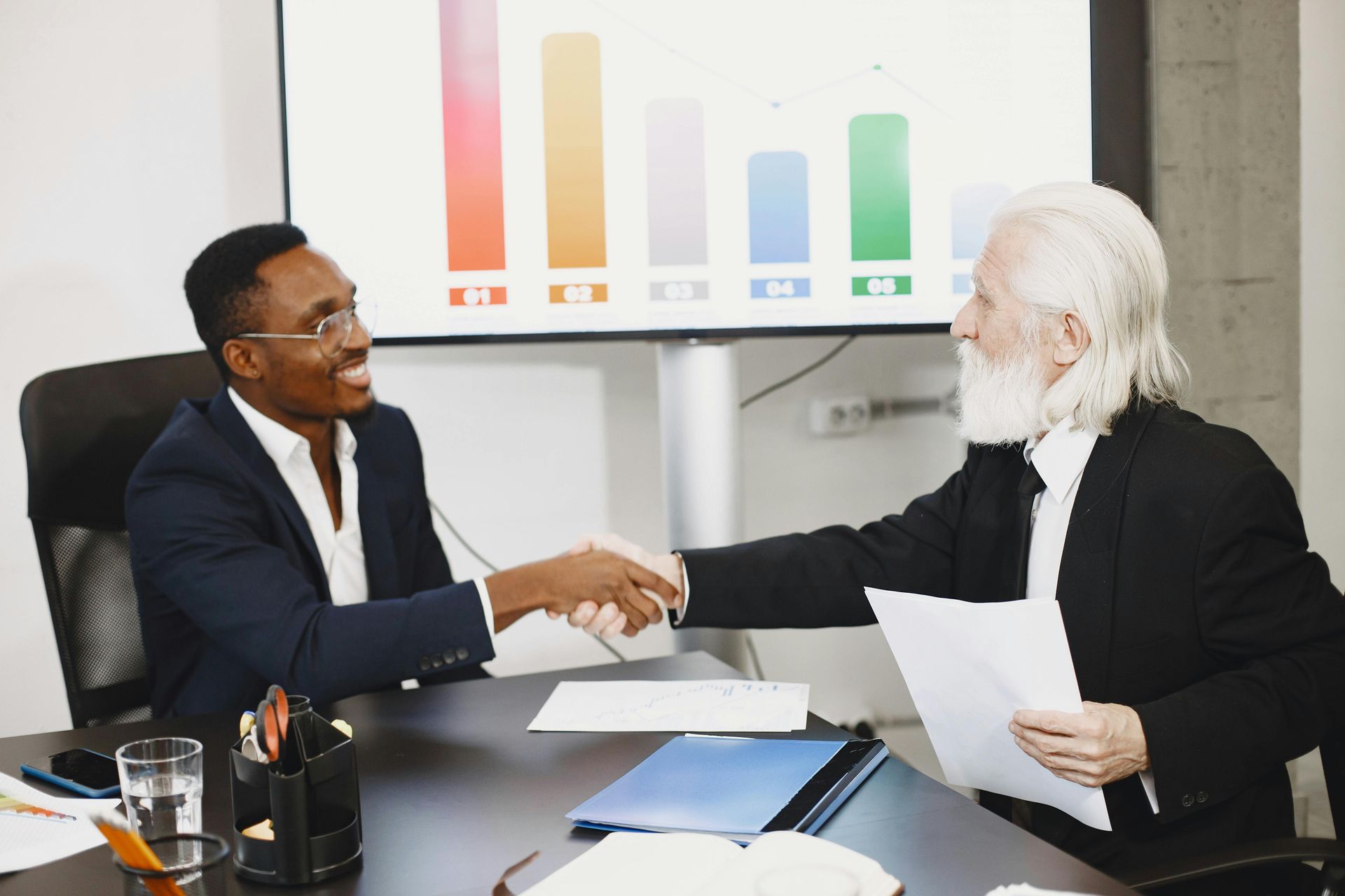 A man is sitting at a table with a laptop and shaking hands with a man.