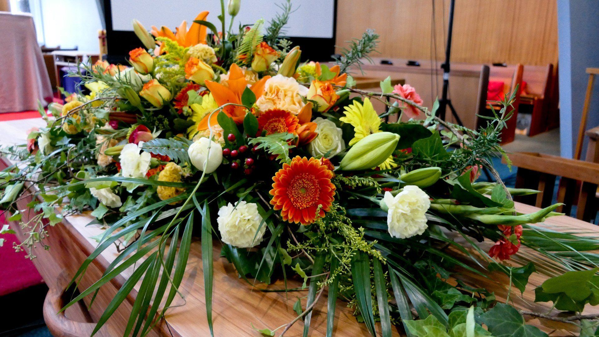 Composizione floreale su una bara di legno in una stanza con schermo per proiettore. I fiori sono arancioni, gialli e bianchi.