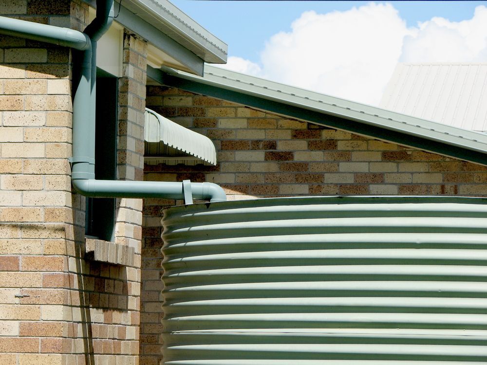 A Corrugated Metal Water Tank Sits Next to a Brick House — Port Macquarie Plumbing In Port Macquarie, NSW