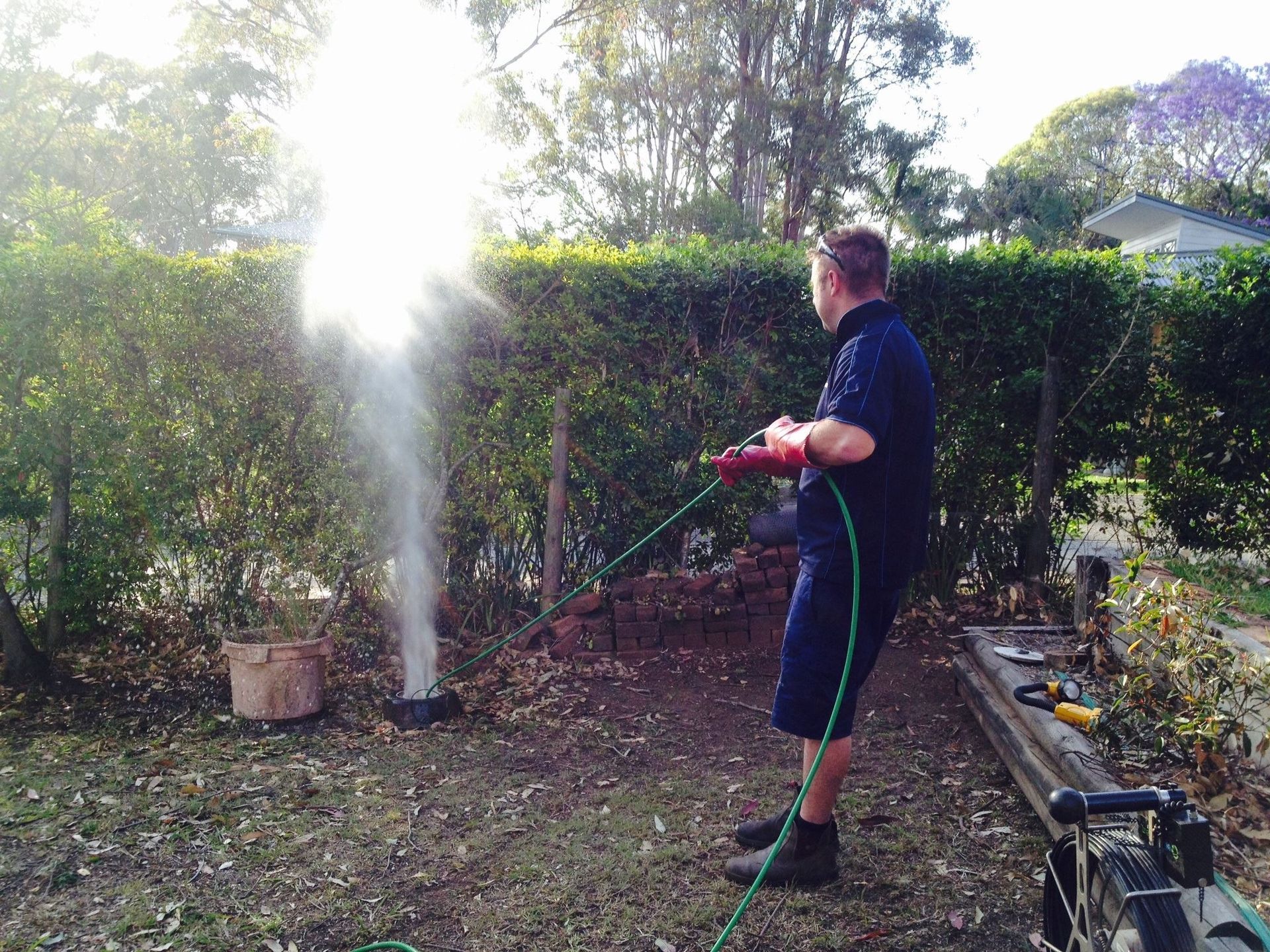 A Person in Uniform Sprays Water from A High Pressure Hose onto A Garden Bed in Front of A Hedge — Port Macquarie Plumbing In Port Macquarie, NSW
