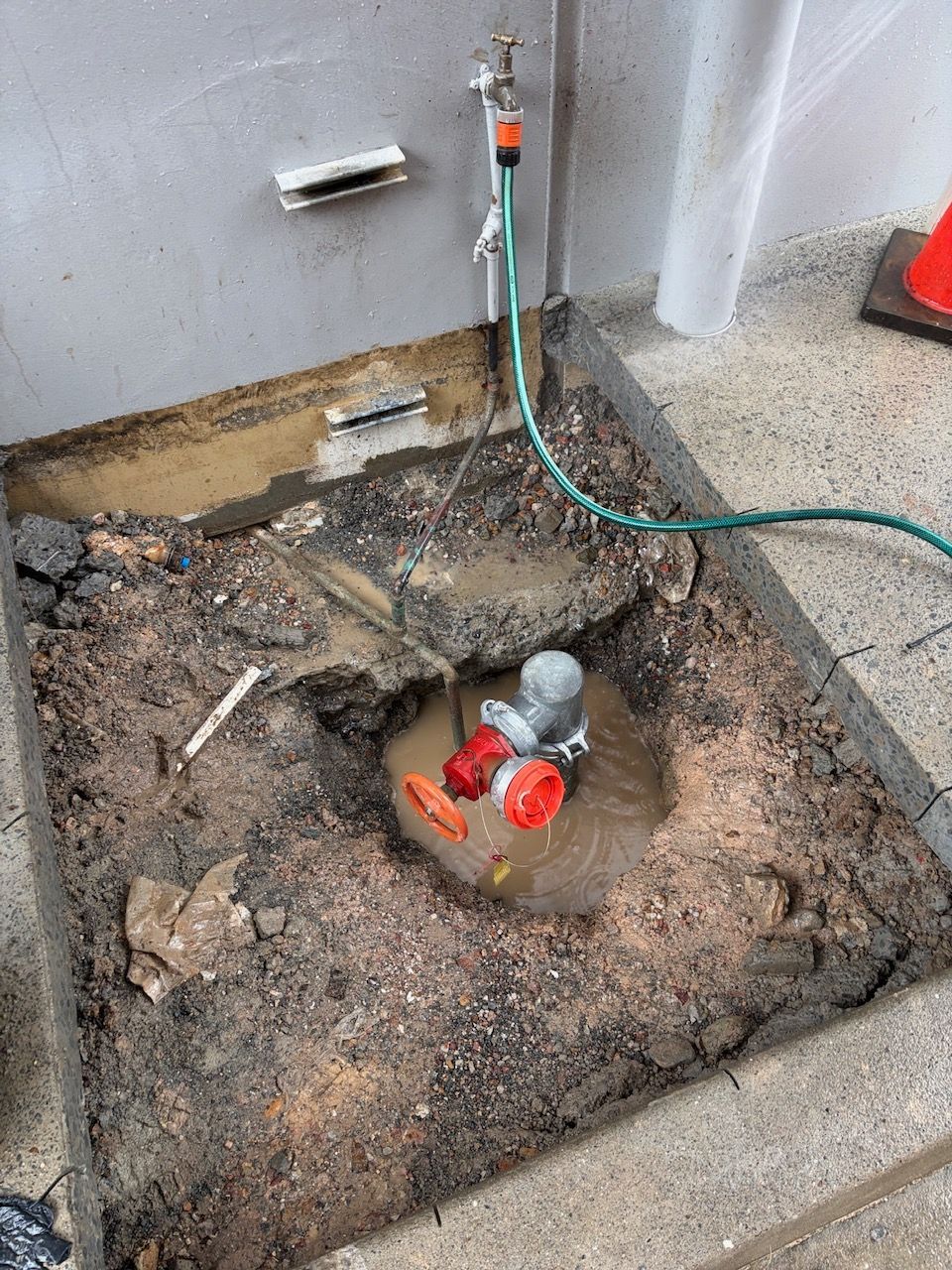 Excavated Ground with A Red Water Meter or Valve and Hose Connected Near a Wall Faucet — Port Macquarie Plumbing In Laurieton, NSW