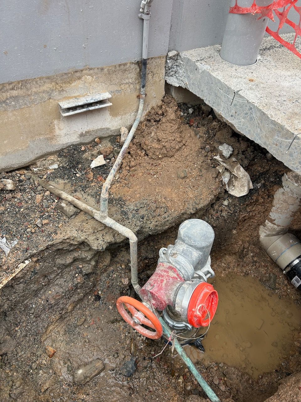 Exposed Underground Water Valve and Pipes in A Muddy Excavation Beside a Concrete Wall — Port Macquarie Plumbing In Laurieton, NSW