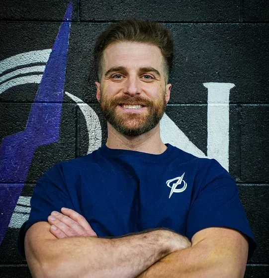 Smiling man in a blue shirt with arms crossed against a dark wall background