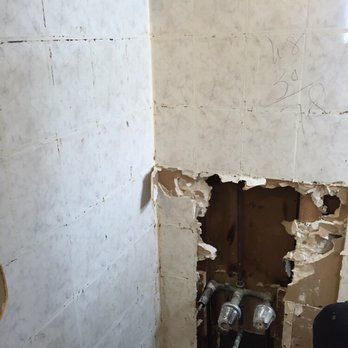 Corner of a damaged wall with exposed plumbing fixtures. Tile is cracked and torn, revealing wood framing.