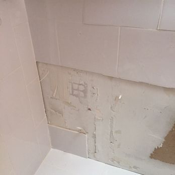 Damaged bathroom wall with missing tiles exposing the underlying drywall and mortar.