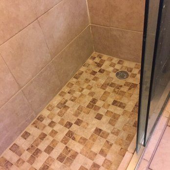 Shower stall with beige tiled walls and mosaic tile floor. Glass door on the right. Drain in floor's center.