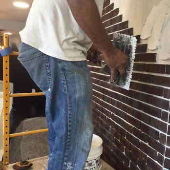 Person applying mortar to a brick wall with a trowel, bucket, and scaffolding nearby.