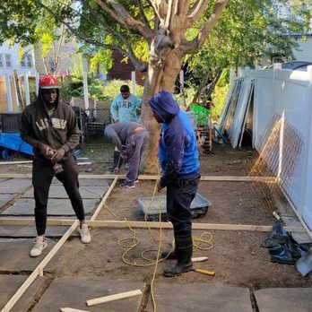 People building a structure in a backyard. One in a red hat watches, others work on wooden framing.